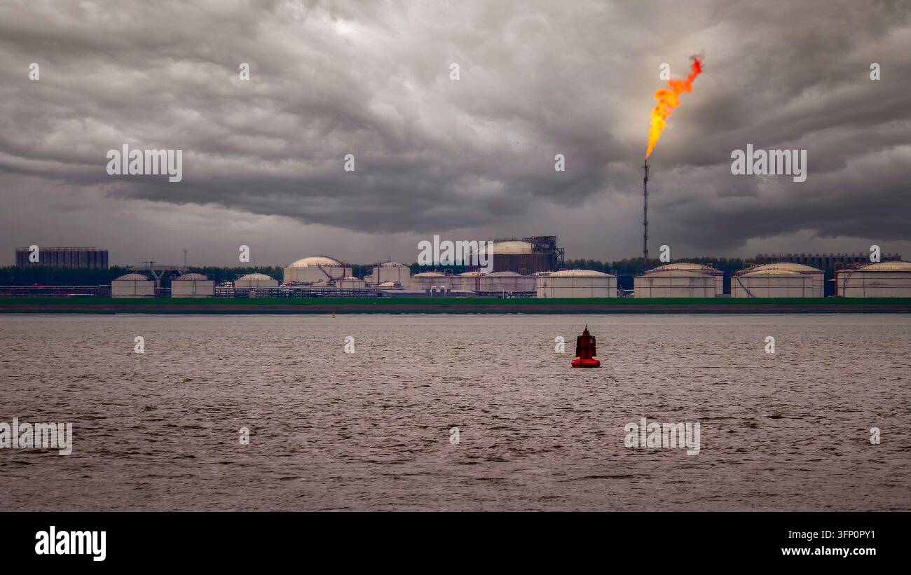 Raffineria industriale di petrolio con serbatoi di stoccaggio e una torcia di fiamma che brucia il gas in eccesso, creando inquinamento sotto un cielo scuro e nuvoloso con una boa in primo piano Foto Stock
