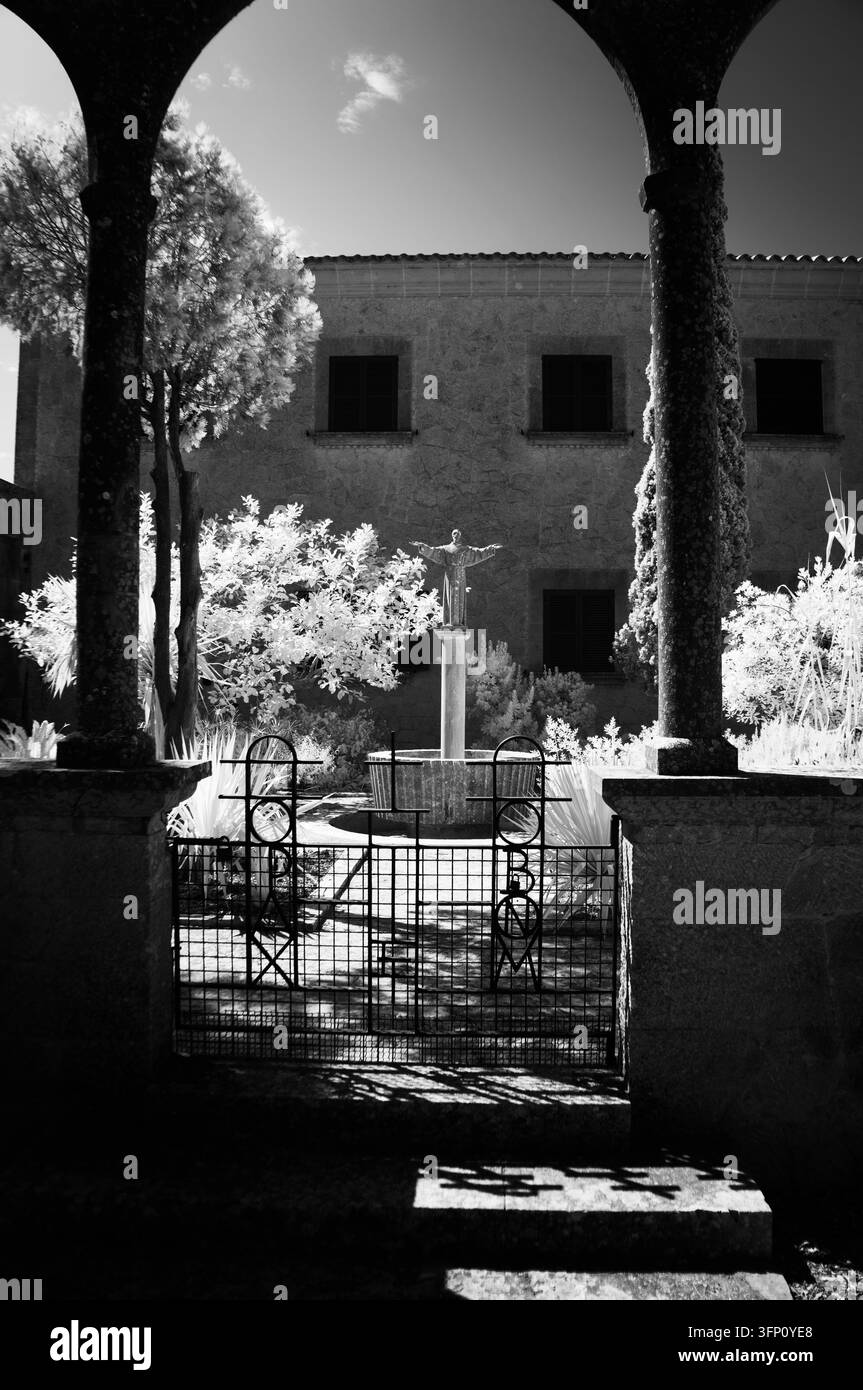 Una fotografia a infrarossi in bianco e nero che mostra un giardino strutturato con una statua religiosa e una fontana, vista attraverso un ingresso ad arco. L'albero Foto Stock