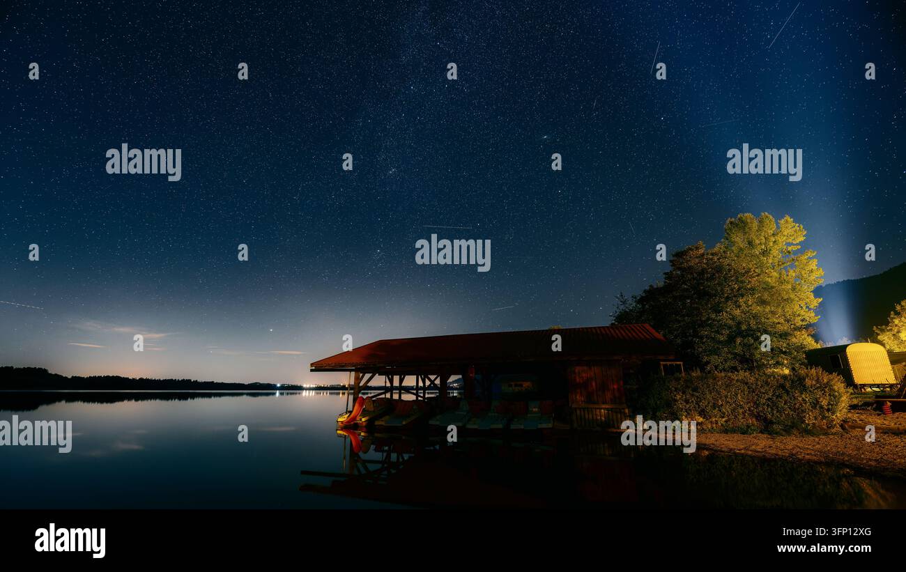 Una rimessa per barche e una fila di alberi illuminati costeggiano la riva del Bannwaldsee sotto un cielo notturno limpido pieno di stelle. Foto Stock