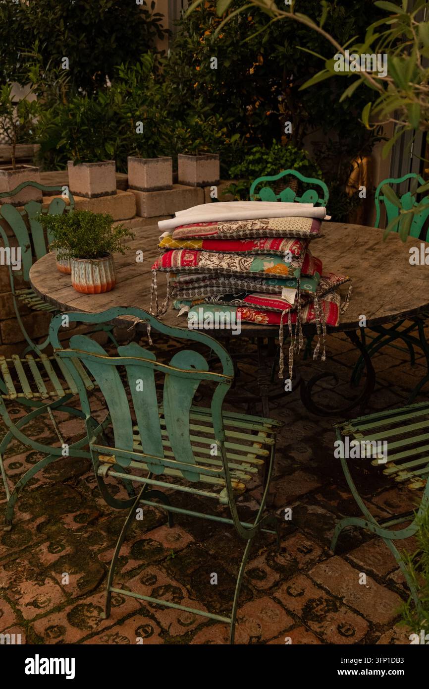 Pila di tessuti colorati piegati su un tavolo rotondo da giardino con sedie in metallo turchesi vintage e piante in vaso su un patio Foto Stock