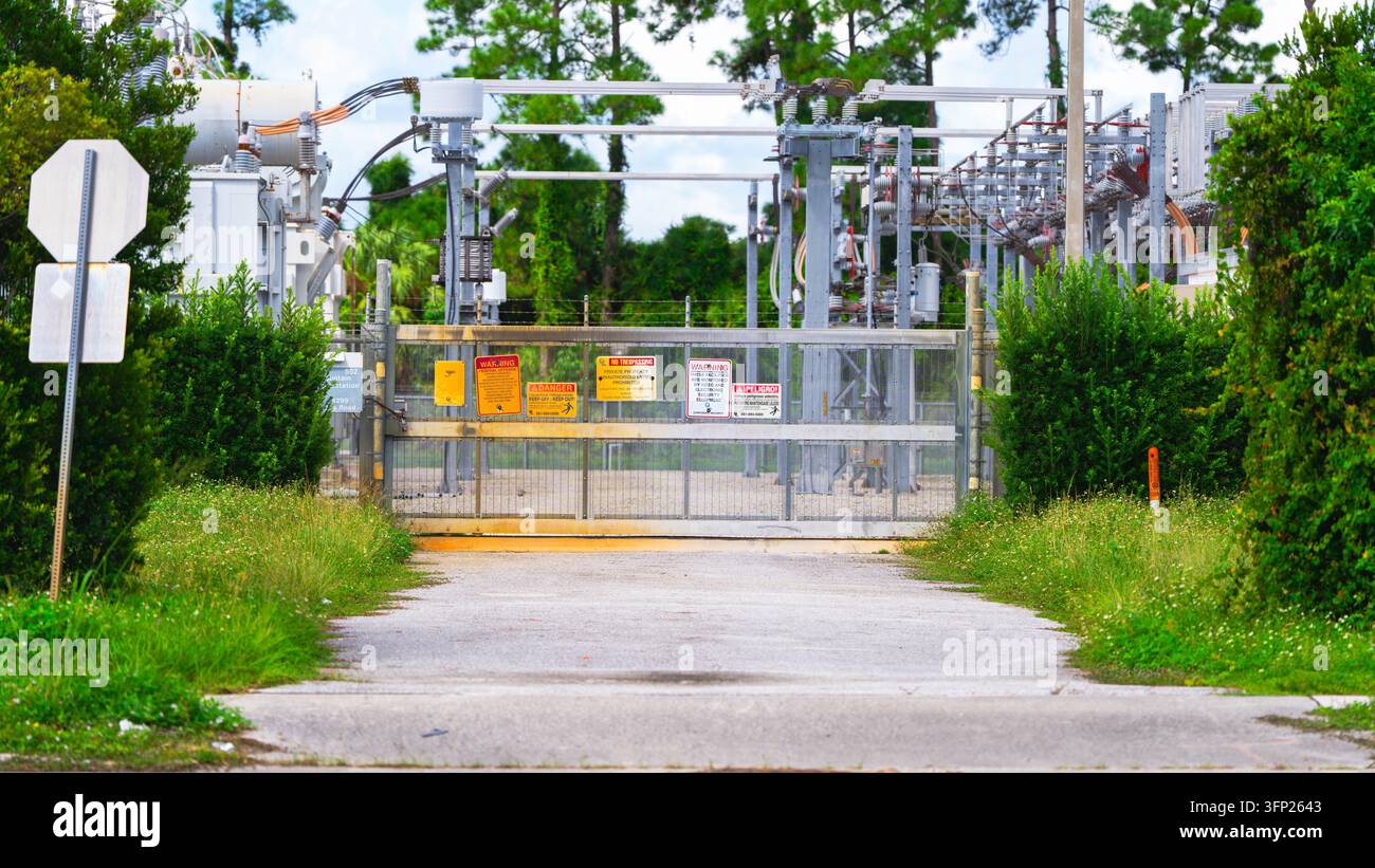 Sottostazione elettrica recintata con segnali di avvertimento e apparecchiature di distribuzione alla fine di una stretta strada di accesso circondata da vegetazione verde in un'area all'aperto. Foto Stock