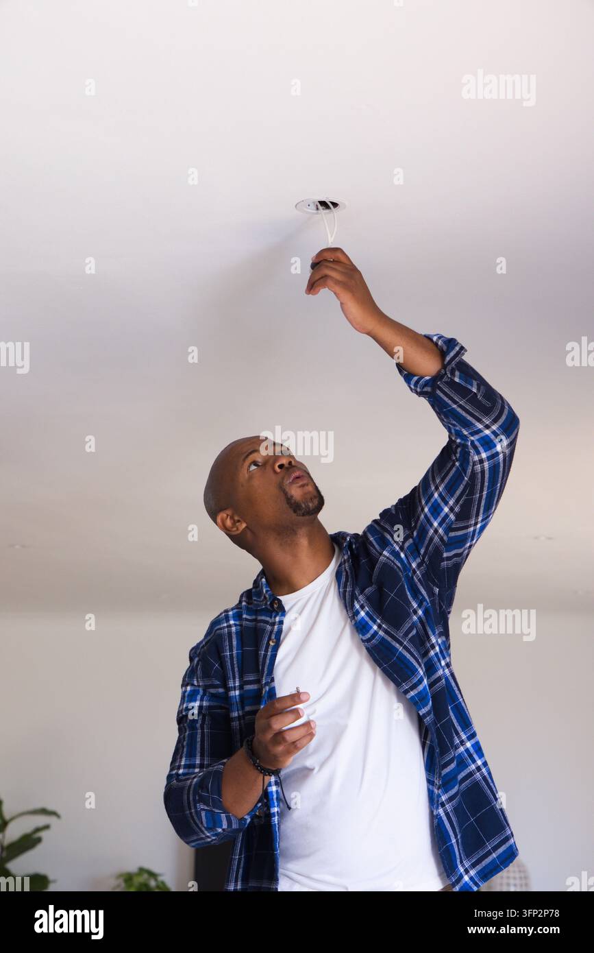 Uomo adulto in piedi sotto un soffitto bianco, indossa una camicia a quadri blu, raggiunge una scatola elettrica aperta Foto Stock
