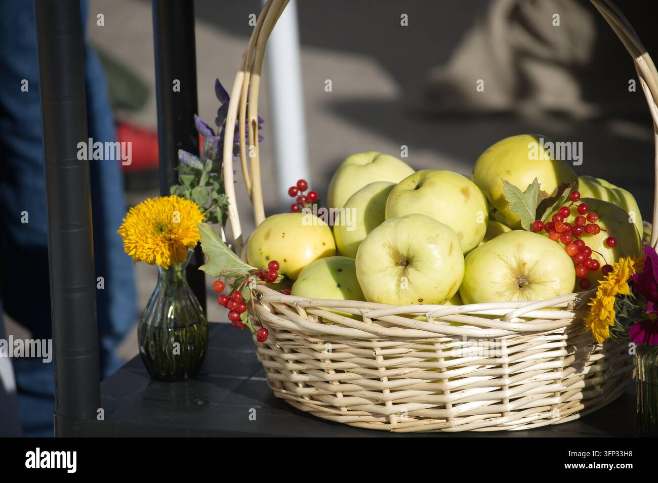 Un contenitore intrecciato rustico contiene frutta di frutteto raccolta accentata da fiori selvatici vibranti in piccoli vasi di vetro durante un luminoso evento di raccolta all'aperto Foto Stock