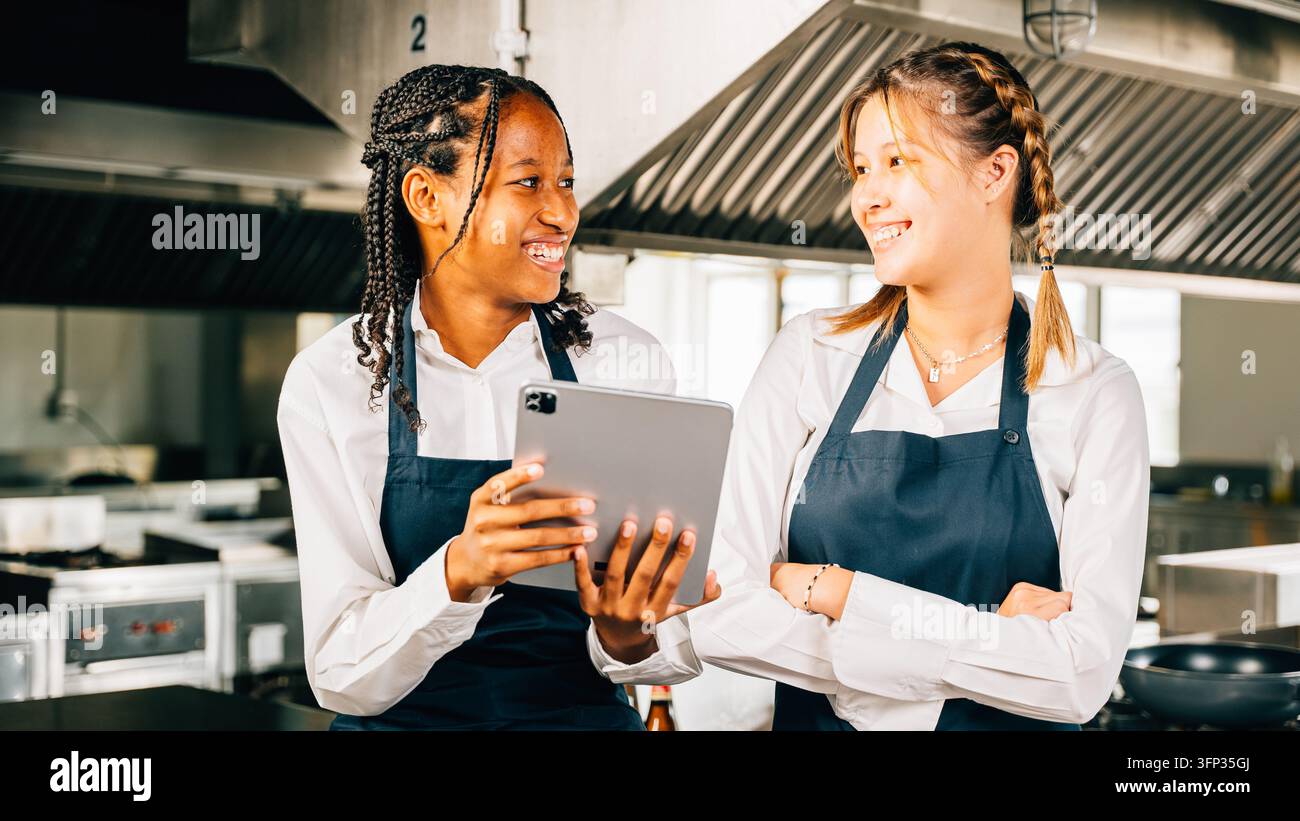 In una grande cucina professionale due famosi chef discutono del video blog usando un tablet. Professionisti sorridenti in piedi leggono cercano la tecnologia moderna a Foto Stock