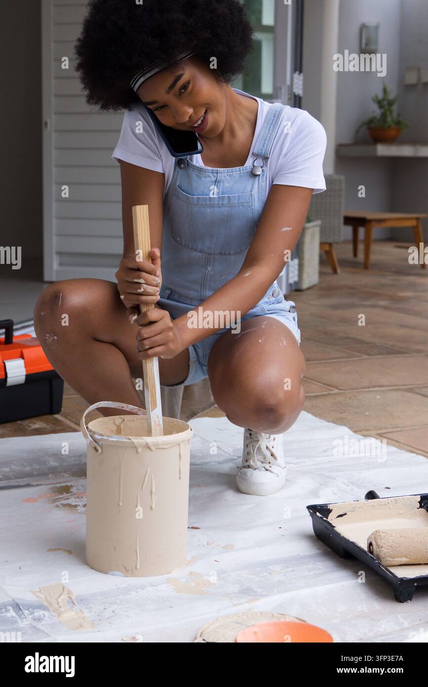 Donna inginocchiata su un telo che mescola la vernice beige con un bastone di legno mentre parla al cellulare Foto Stock