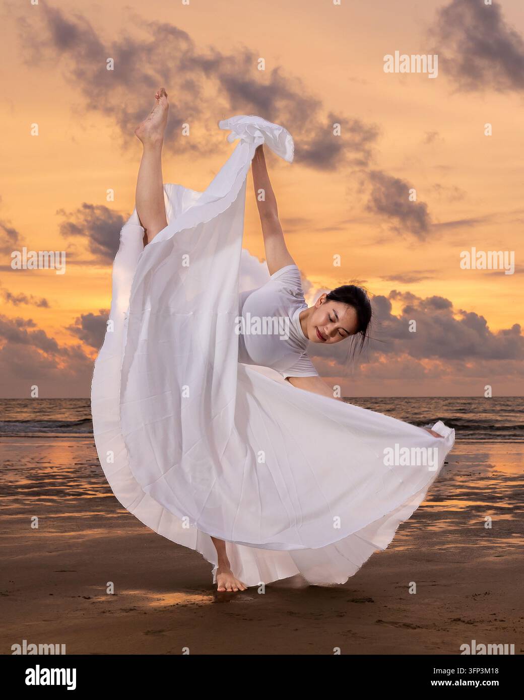 Ginnasta femminile che esegue una posa ginnica elegante e flessibile in un vestito bianco fluente su una spiaggia durante un tramonto dorato Foto Stock