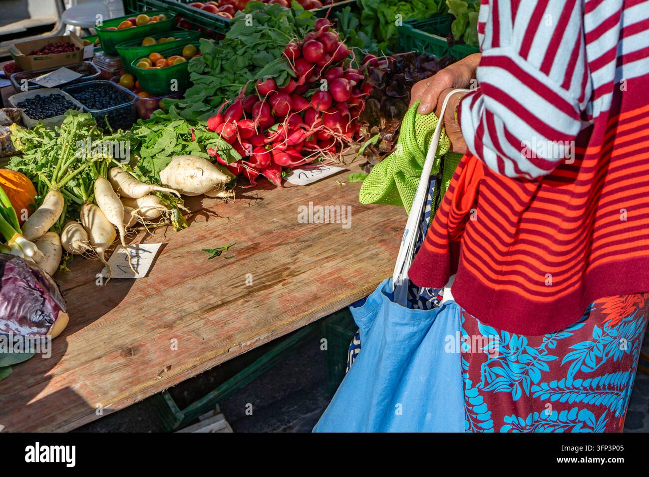 Una donna anziana che vende ortaggi freschi in un mercato locale. I prodotti stagionali sono esposti al suo banco, evidenziando le vendite tradizionali di cibo Foto Stock