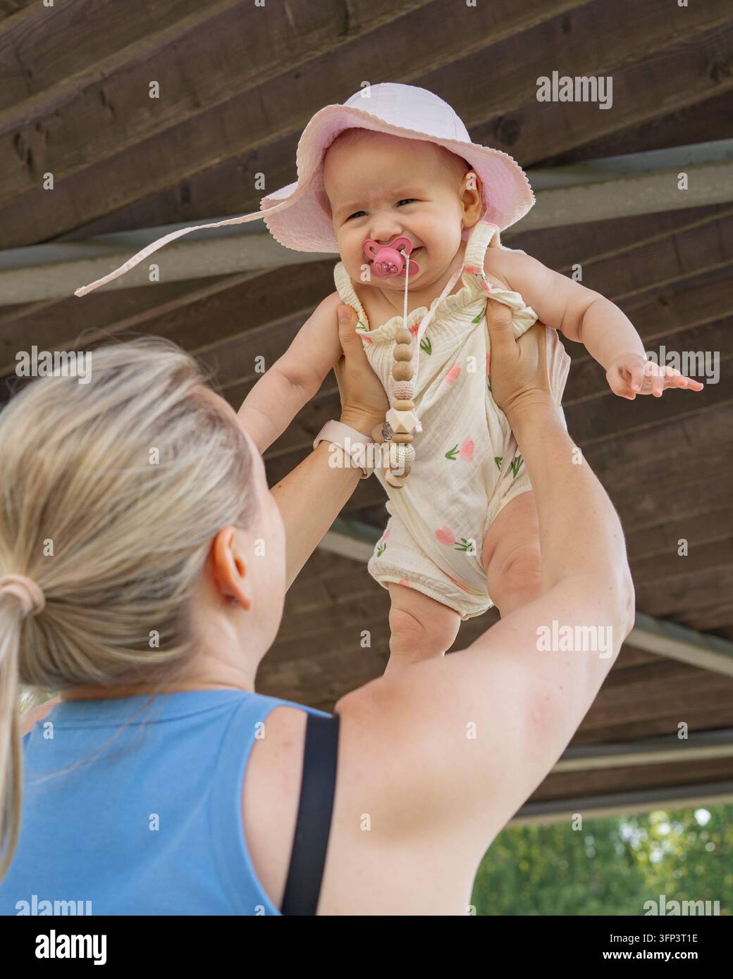 Un bambino gioioso sollevato sopra la spalla da una donna in una struttura di legno all'aperto, circondato da abiti rosa e bianchi e una collana di perle Foto Stock