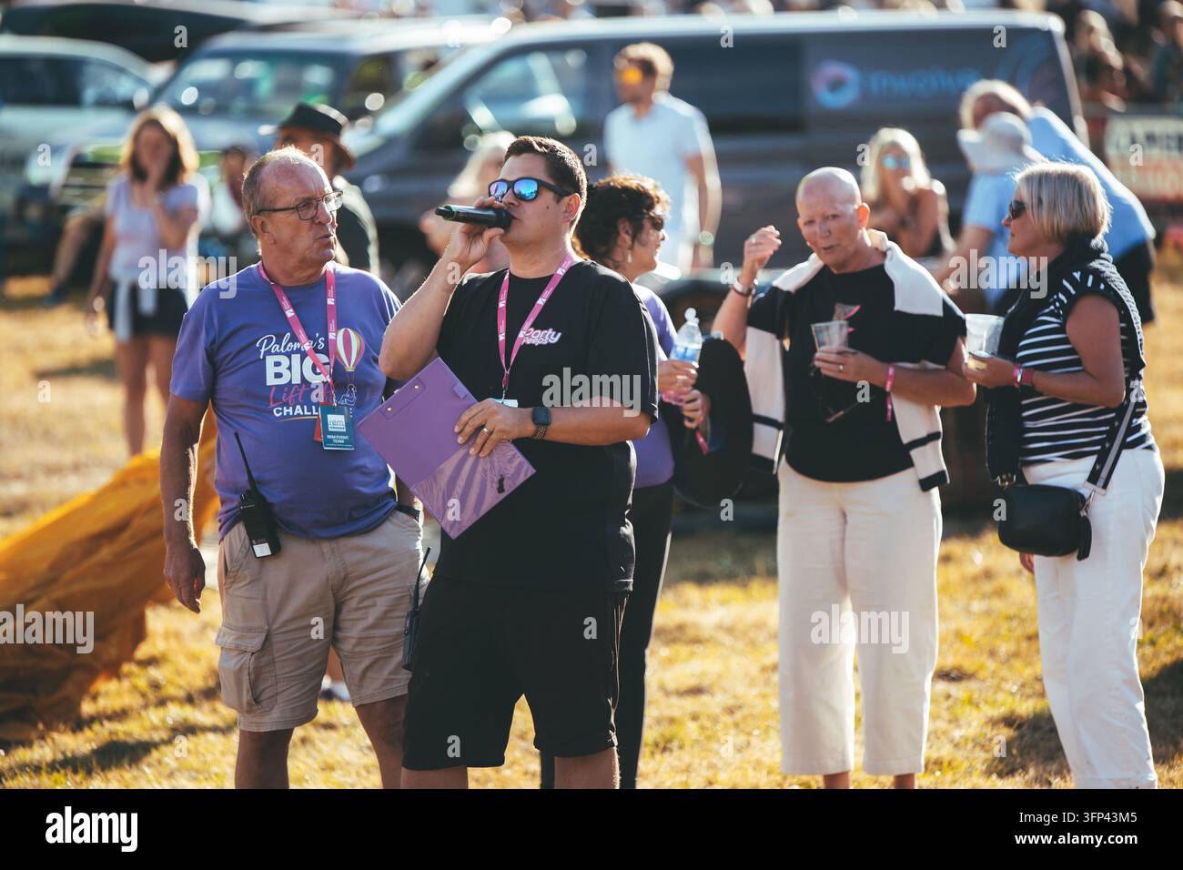 Bristol Balloon Fiesta, 7 agosto 2026, UK – Membro del personale dell’evento che parla in un microfono accanto all’annunciatore e ai partecipanti su un campo festival soleggiato Foto Stock