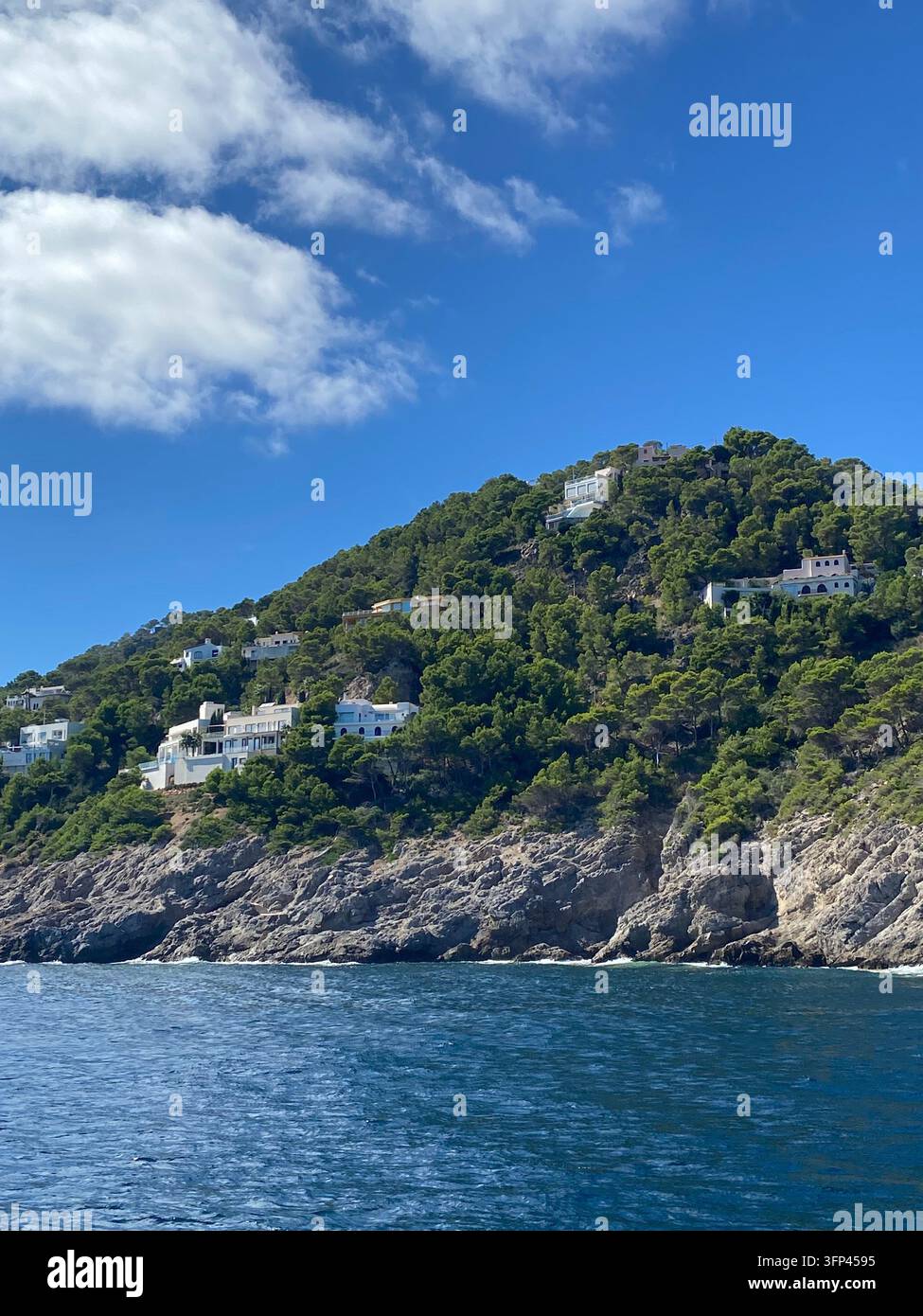 Vista scenica dal mare di una collina lussureggiante con ville bianche di lusso arroccate su una costa rocciosa sotto un cielo blu brillante con nuvole. - Immagine stock catturata con smartphone
