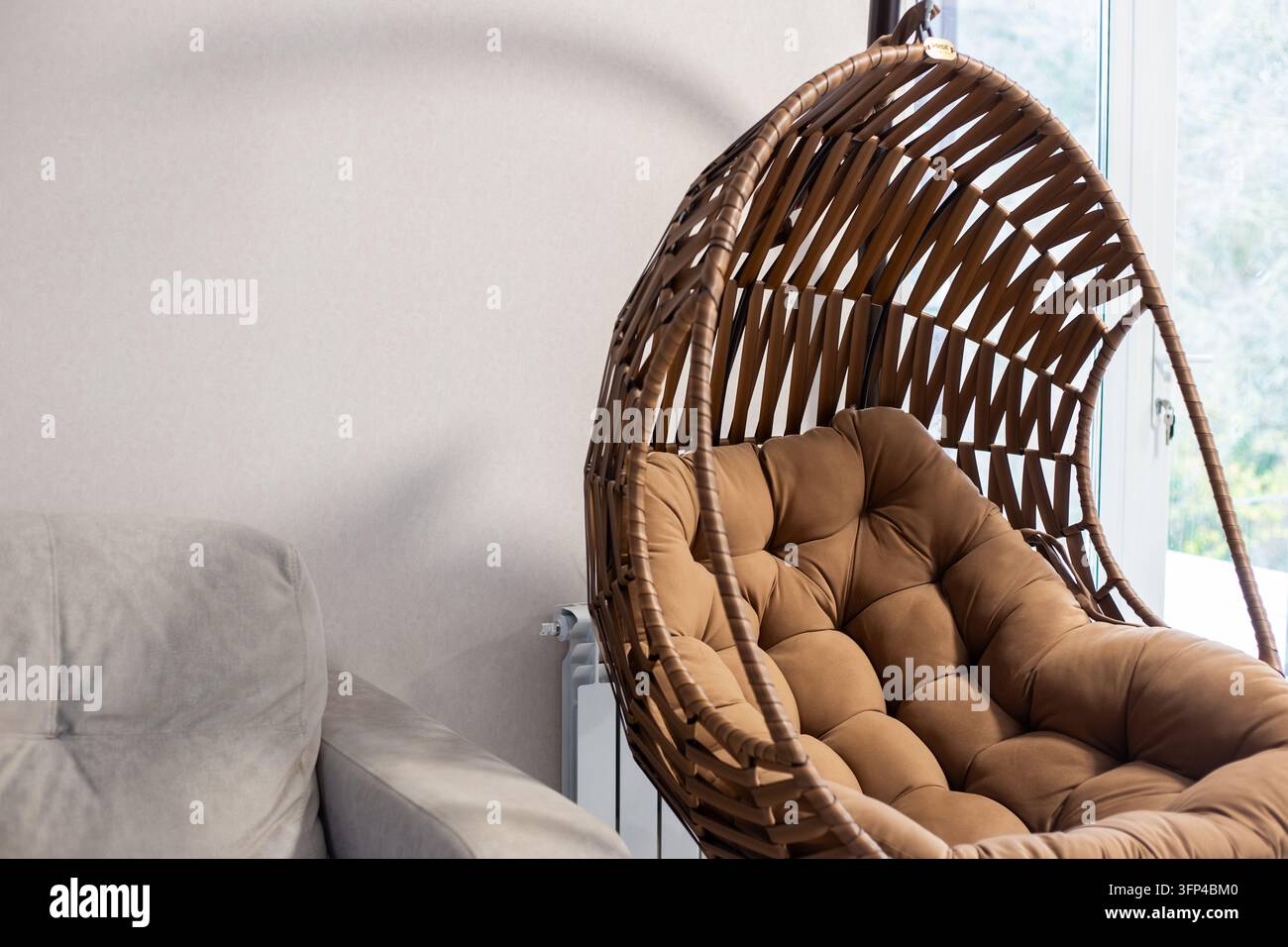 Un'elegante sedia a uovo sospesa in vimini marrone con un morbido cuscino si trova accanto a un divano. Disposizione di design di interni di appartamento moderno e accogliente. Tema di mobili. Foto Stock