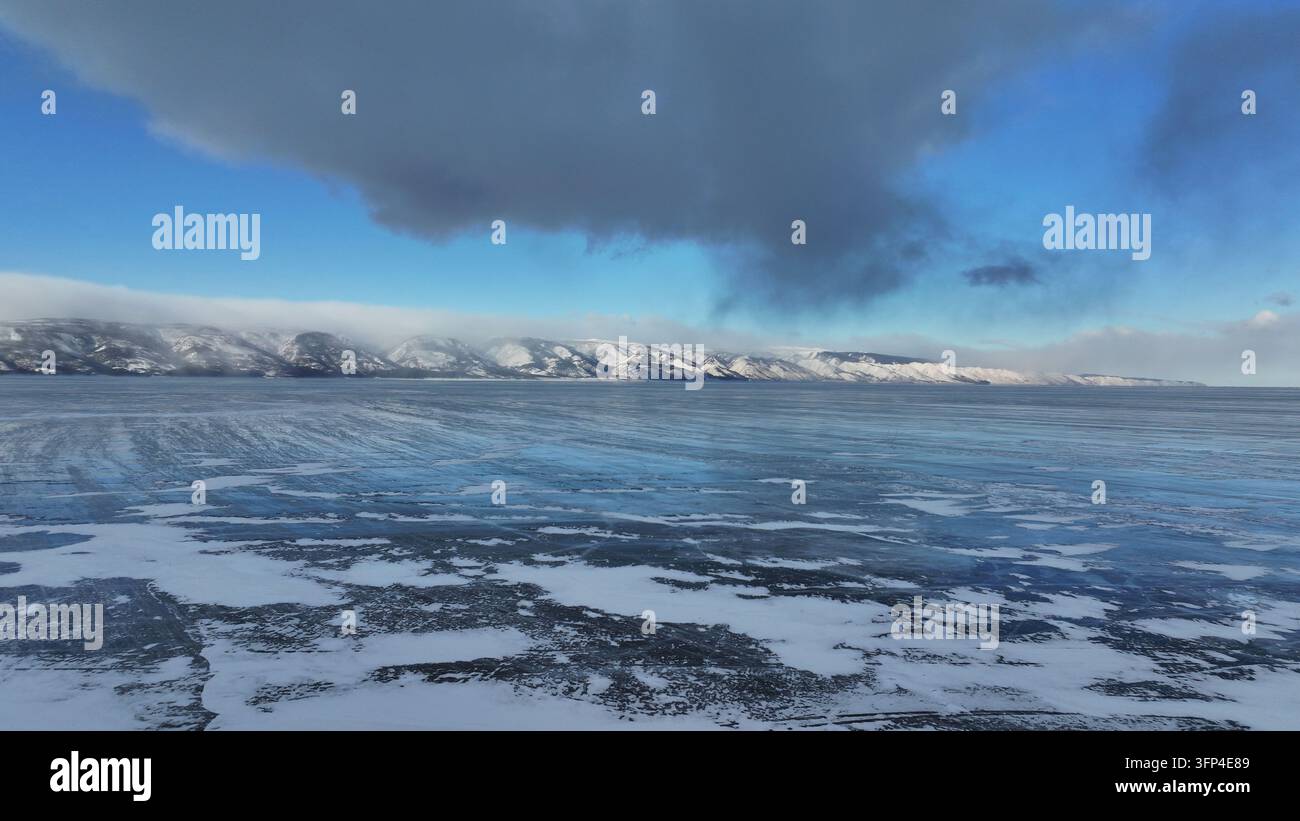 Un panorama aereo si estende sul ghiaccio e sulla neve irregolari del lago Baikal verso una lunga fila di montagne innevate. Nuvole invernali drammatiche incombono sul re Foto Stock