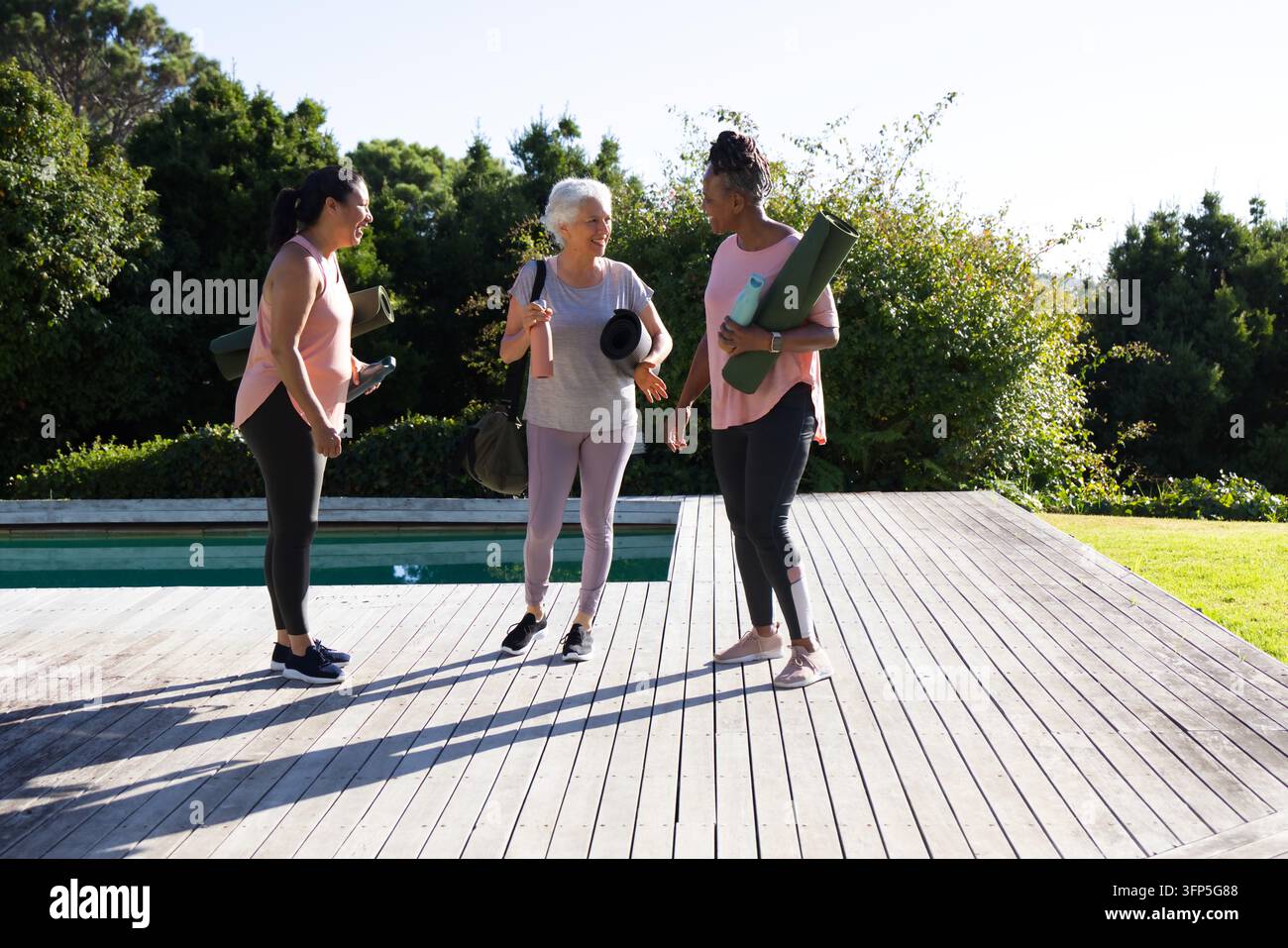 Diverse amiche in abbigliamento sportivo in piedi su un deck di legno vicino alla piscina con materassini da yoga Foto Stock