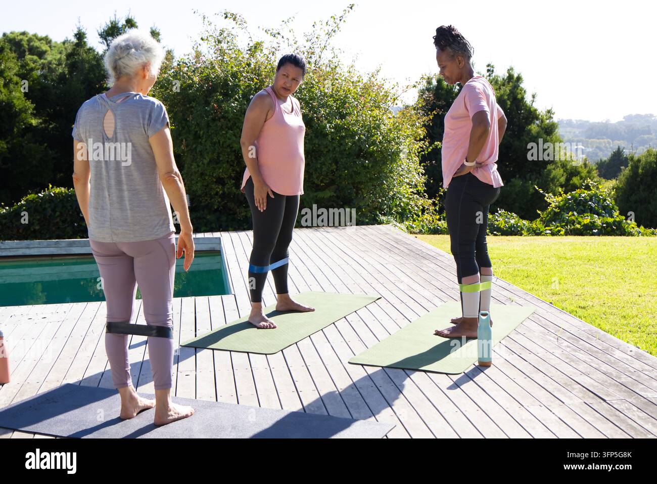 Diverse amiche che fanno esercizio su materassini verdi su un terrazzo di legno vicino alla piscina indossando abbigliamento sportivo Foto Stock