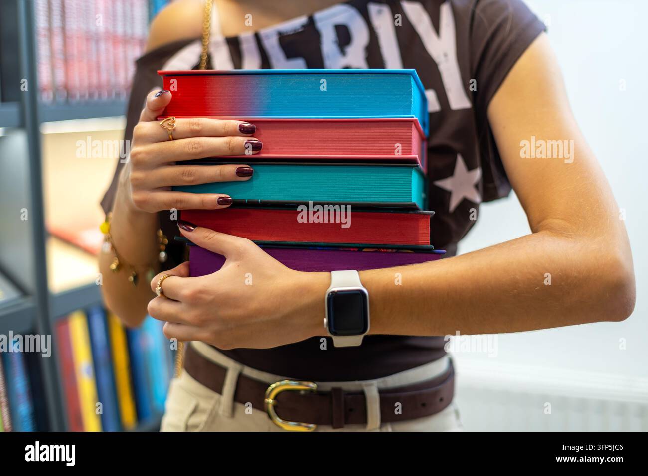 Giovane donna che tiene una pila di libri colorati in biblioteca, studente che porta materiale di studio per l'istruzione, lettura e apprendimento accademico. Foto Stock