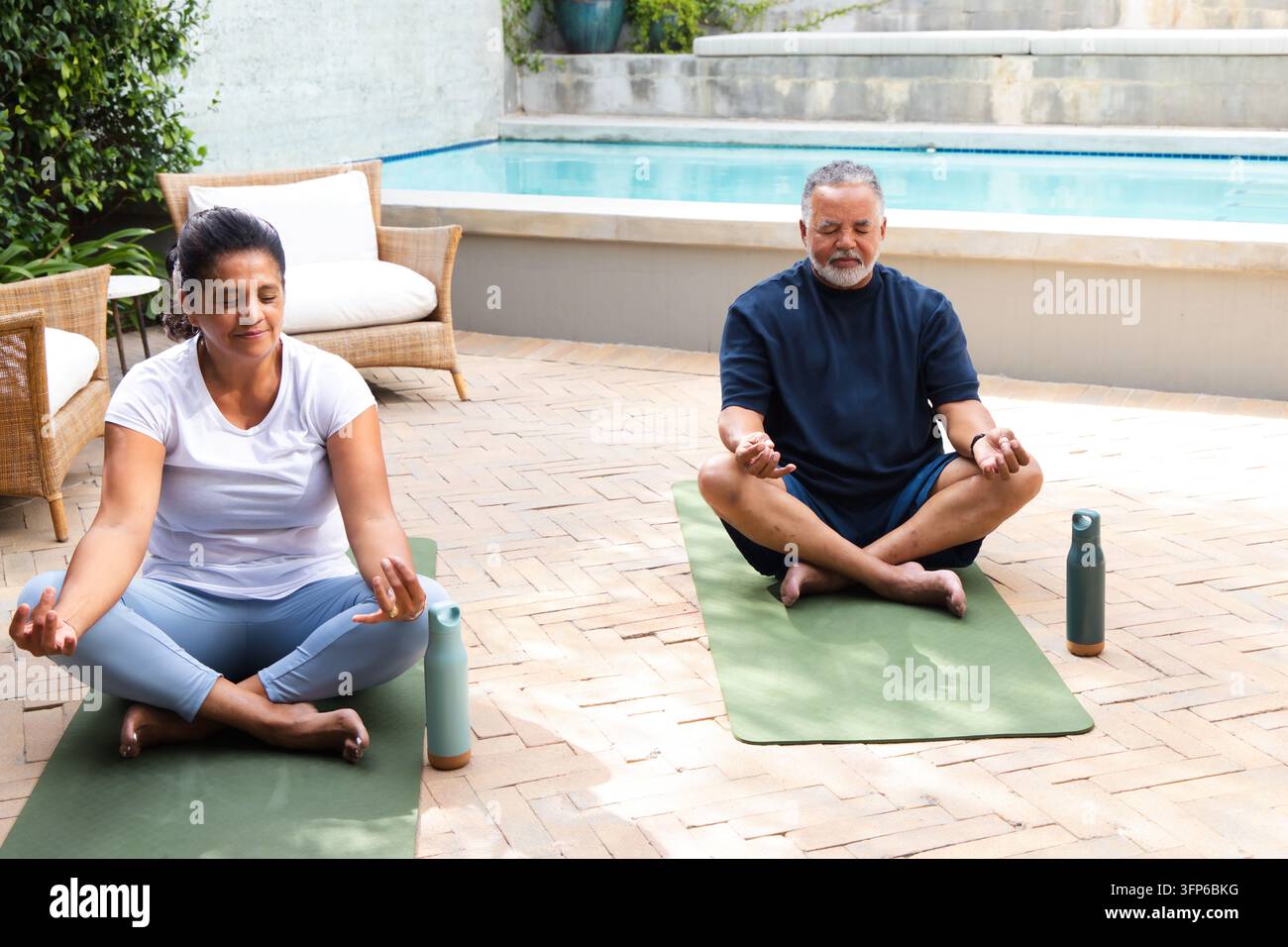Coppia di anziani che medita a gambe incrociate su tappetini da yoga verdi in un patio vicino alla piscina accanto a bottiglie d'acqua Foto Stock