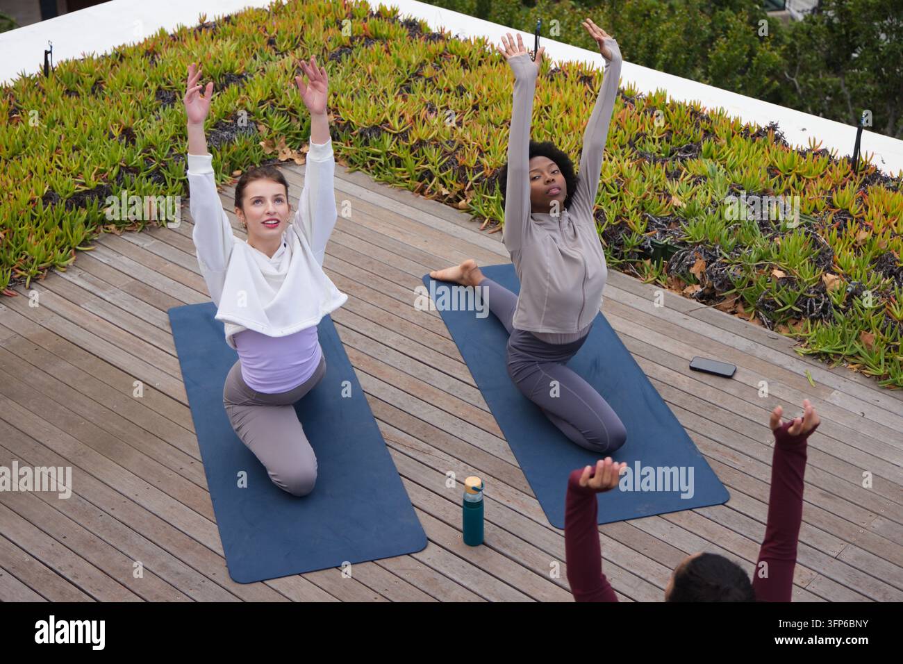 Diverse donne in abbigliamento sportivo che praticano yoga in ginocchio su un terrazzo sul tetto con materassini blu, bottiglia d'acqua Foto Stock