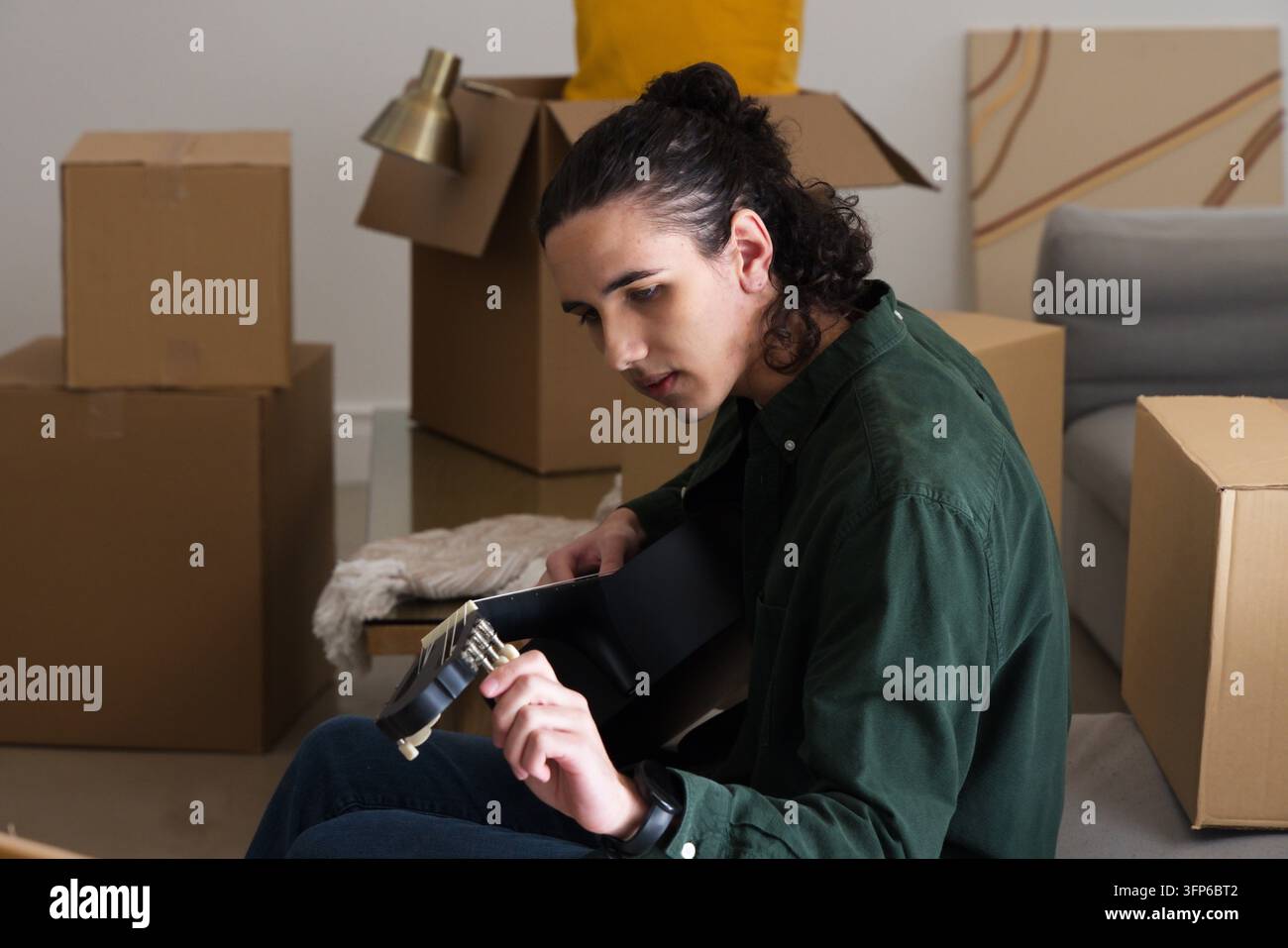 Uomo con camicia verde scuro seduto sul pavimento che accorda una chitarra acustica nera tra scatole in movimento Foto Stock