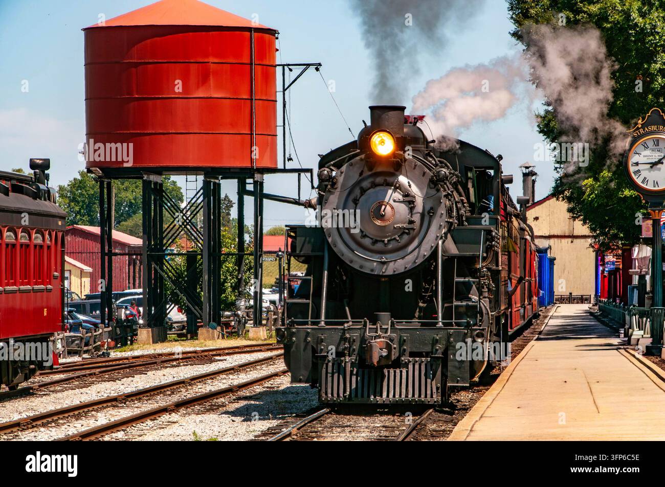 Una locomotiva a vapore entra nella stazione mentre i passeggeri attendono con impazienza il suo arrivo. Il sole splende luminoso, creando un'atmosfera vivace con le persone Foto Stock