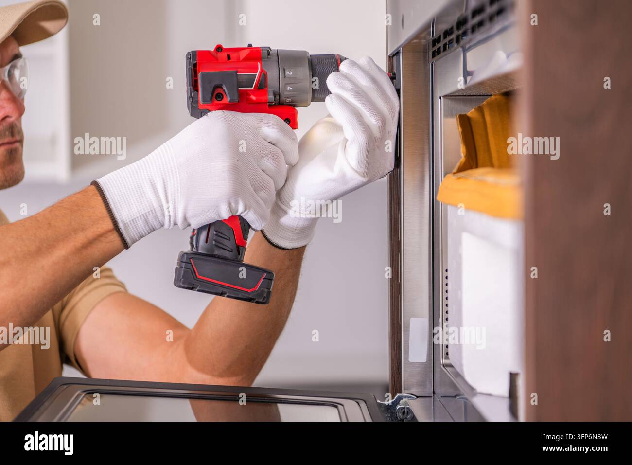 Prima di un tecnico che utilizza un avvitatore a batteria per stringere una vite di fissaggio su un forno incassato all'interno dei mobili della cucina. Foto Stock