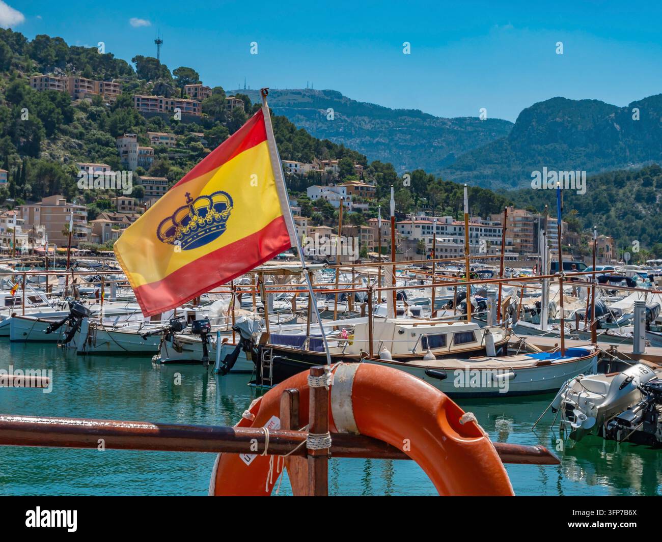 Bandiera spagnola con corona reale sventola sopra il pittoresco porto di pesca di Port de Soller, Maiorca, con tradizionali barche mediterranee di nuovo ormeggiate Foto Stock