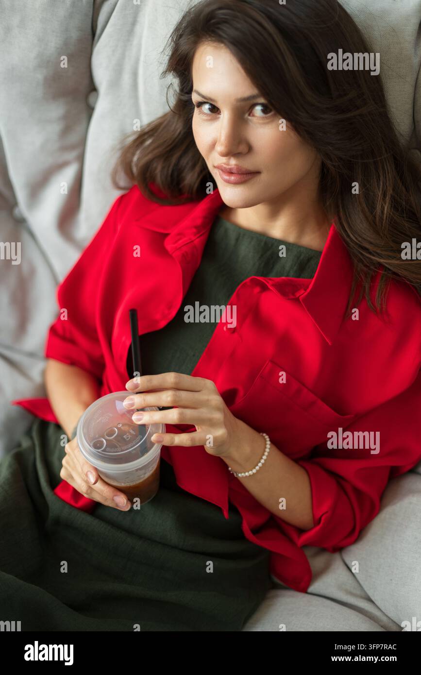 Ritratto fresco di una giovane donna attraente in una camicia rosso acceso che tiene un caffè freddo, guarda direttamente la fotocamera con un sorriso sottile mentre si rilassa Foto Stock