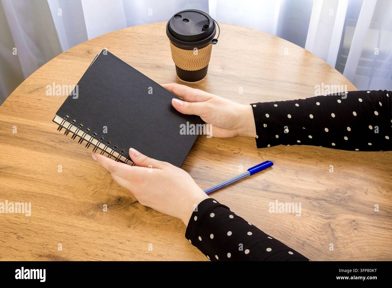 Una donna tiene un quaderno rilegato a spirale nero. Caffè in una tazza monouso è posato su un tavolo nelle vicinanze. Un ambiente domestico con luce naturale. Foto Stock