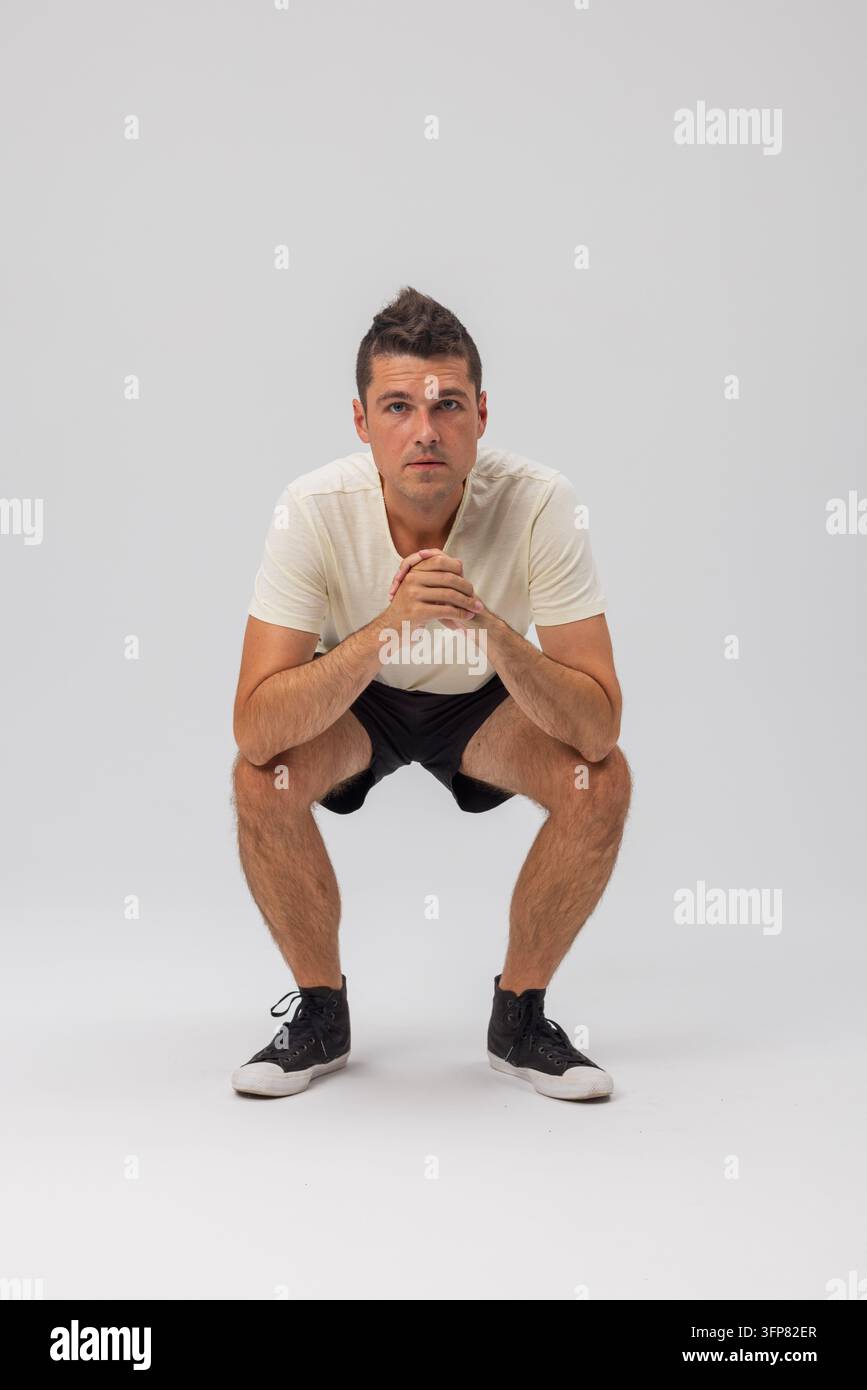 Un giovane uomo si accovaccia su uno sfondo grigio neutro, guardando la fotocamera con le mani giunte. Indossa una maglietta bianca e dei pantaloncini neri. Scarpa da studio pulita Foto Stock