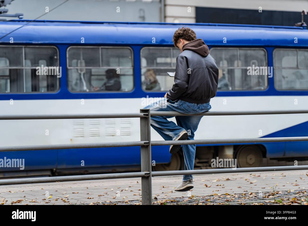 Una persona appollaiata su una barriera pedonale controlla un dispositivo portatile accanto a una tramvia con passeggeri, mostrando l’attività digitale quotidiana all’interno di un pub urbano Foto Stock