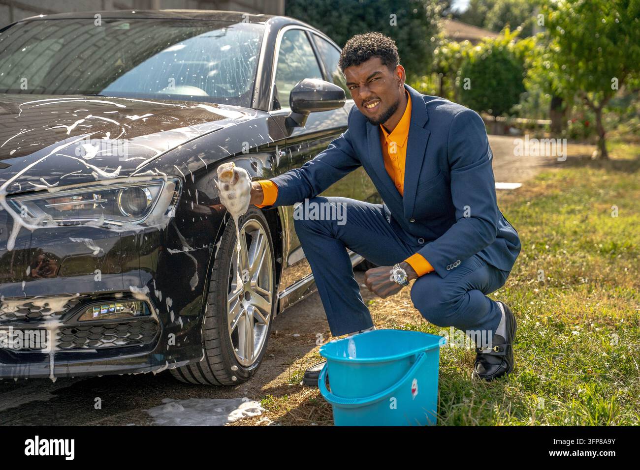 Giovane uomo in completo blu su misura accovacciato accanto a una berlina nera mentre lava la carrozzeria con una spessa schiuma di sapone, blu nelle vicinanze, luce solare brillante sul prato dr Foto Stock
