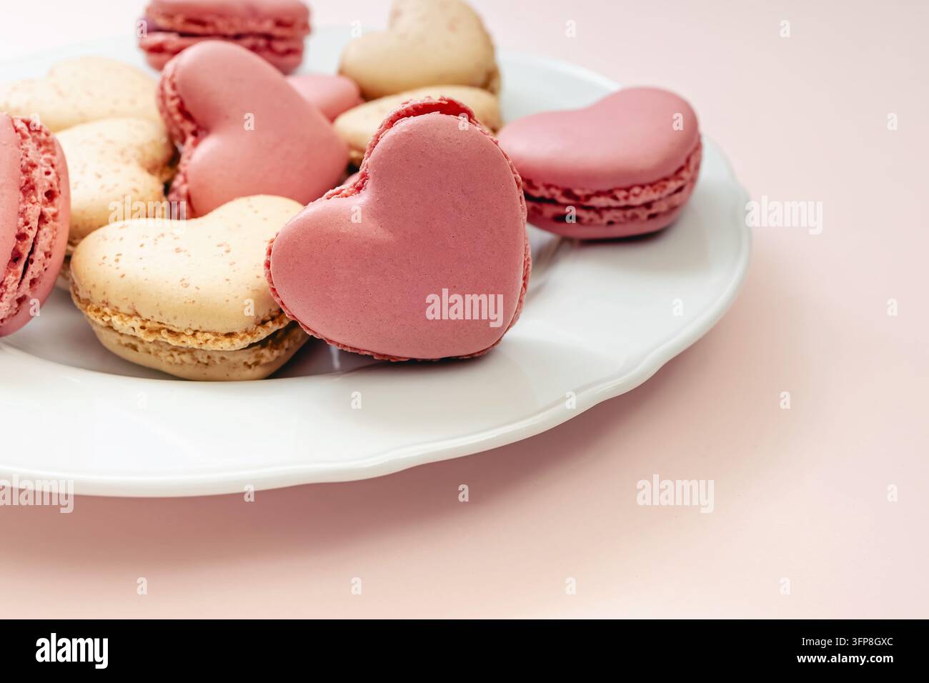 Prima piano di macarons a forma di cuore rosa e vaniglia su un piatto bianco smussato, messa a fuoco su un cuore rosa, sfondo rosa pallido. Foto Stock