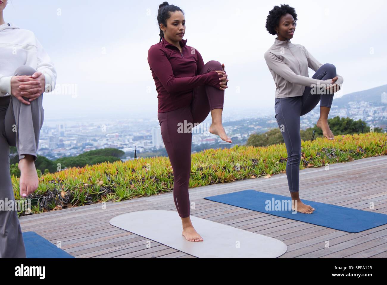 Diverse amiche che praticano l'equilibrio in piedi tenendo il ginocchio al petto su un terrazzo di legno, tappetini da yoga Foto Stock