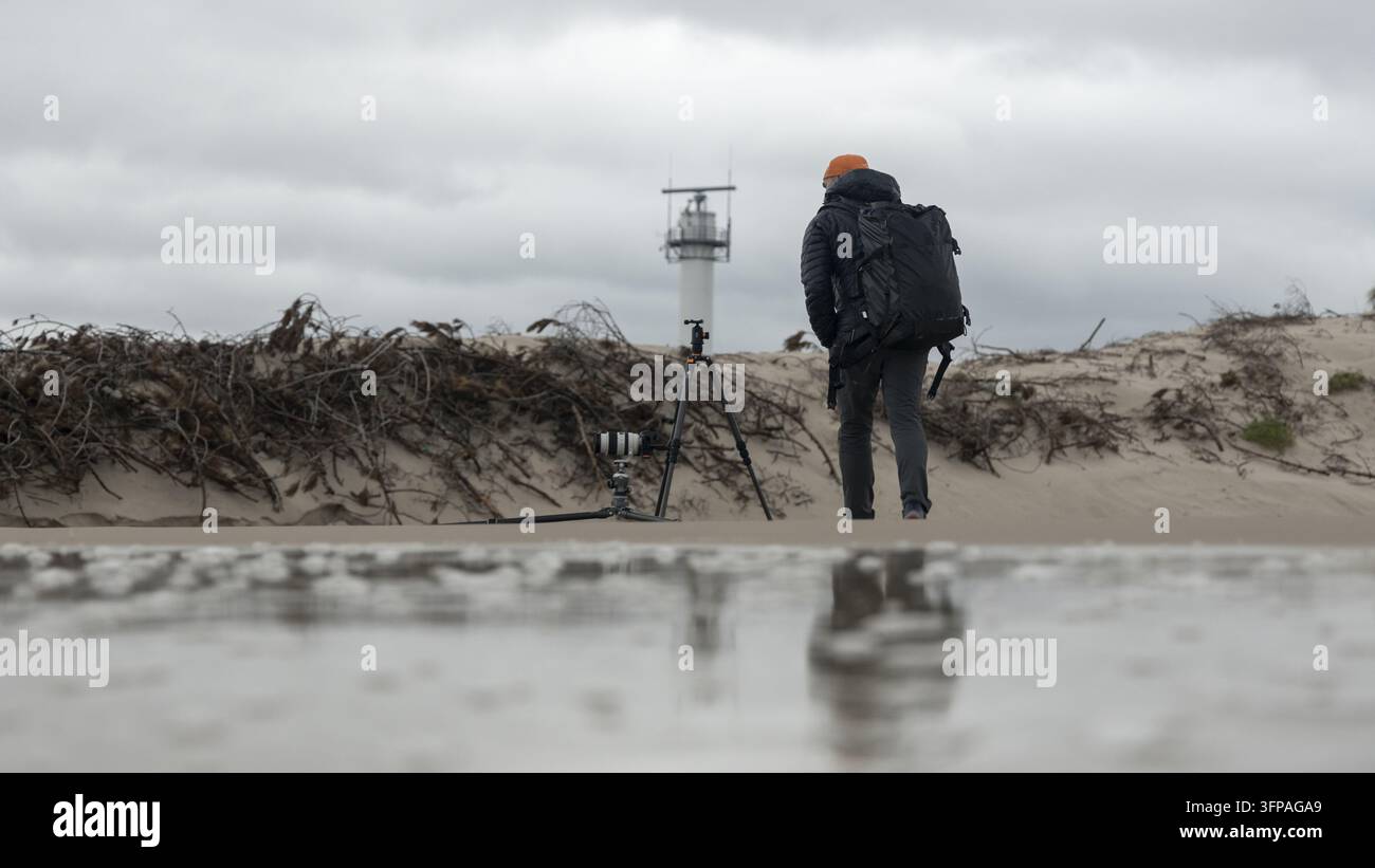 Vista di un fotografo con uno zaino e un treppiede in piedi sulla spiaggia sabbiosa con un faro in lontananza sotto un cielo nuvoloso a Debki, Pomerania Foto Stock