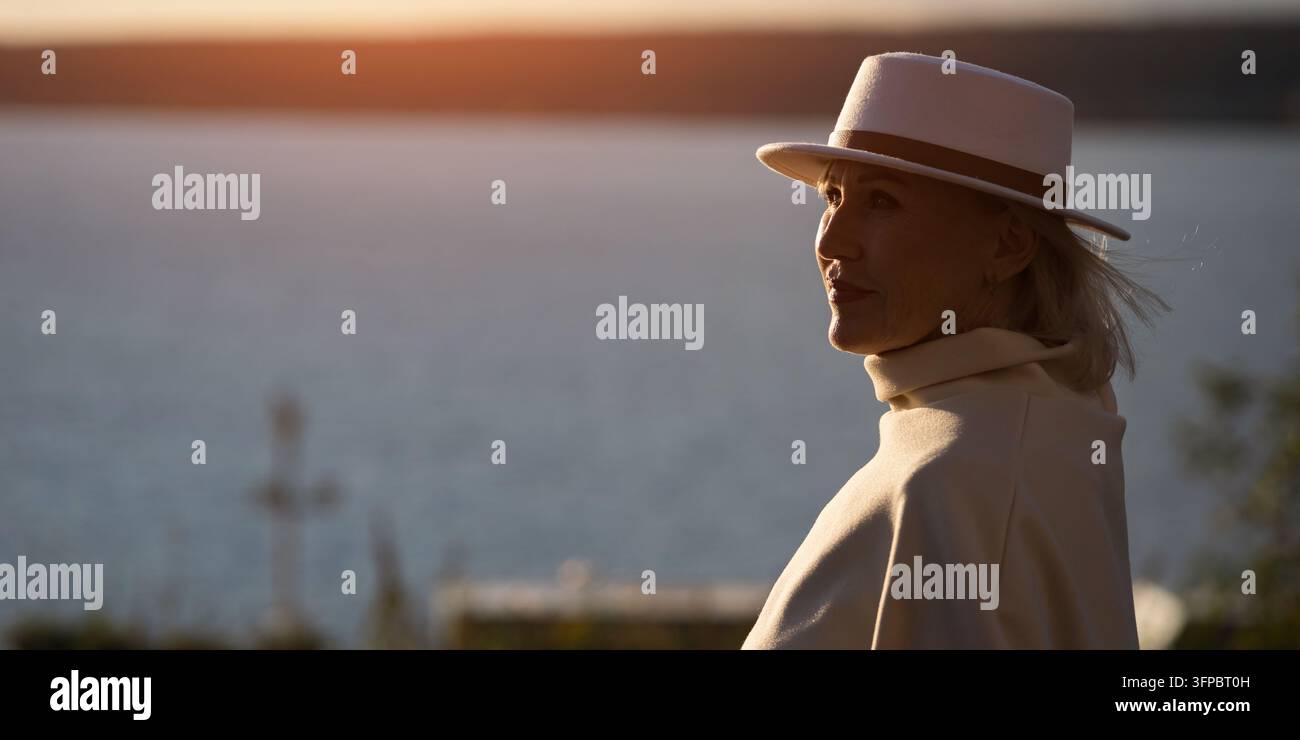 Donna con cappello e cappotto bianco che guarda sull'acqua durante l'ora dorata, trasmettendo calma sicurezza. Viaggiatrice elegante in piedi accanto al lungomare, presentando Foto Stock