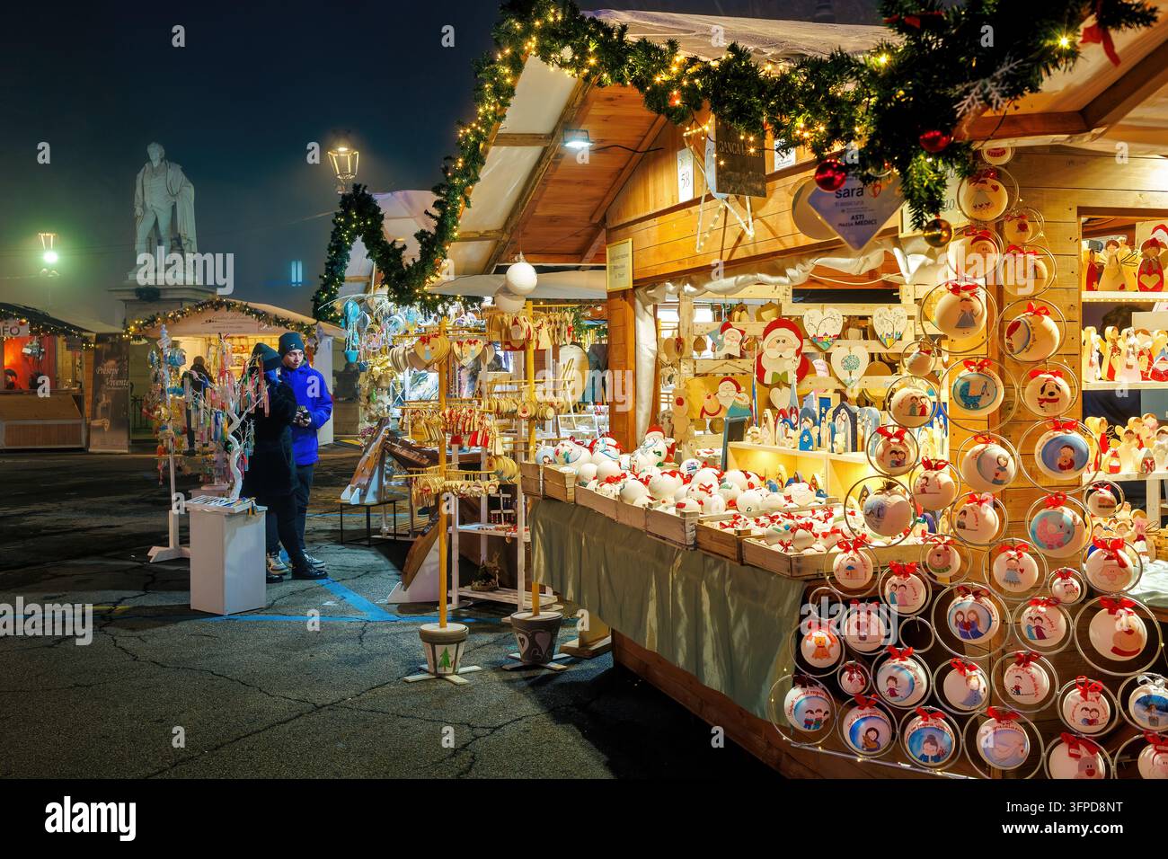 Bancarella festosa del mercato natalizio piena di ornamenti natalizi fatti a mano, decorazioni e luci calde brillanti di notte ad Asti, Italia Foto Stock