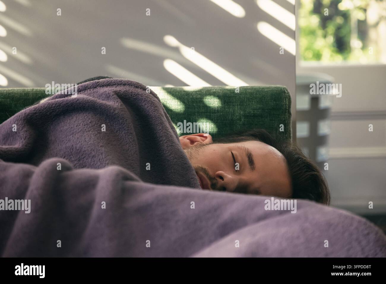 Un uomo caucasico che fa un pisolino pomeridiano sul divano a casa, con la luce del sole che filtra attraverso le persiane Foto Stock