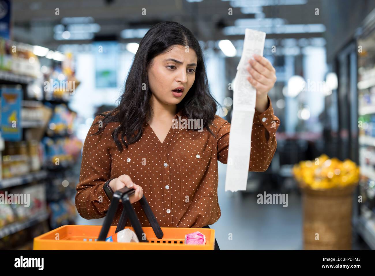 Donna che tiene un lungo scontrino della spesa ed esprime shock e incredulità mentre è in piedi in un corridoio del supermercato, riflettendo l'aumento dei costi e le preoccupazioni sul budget Foto Stock