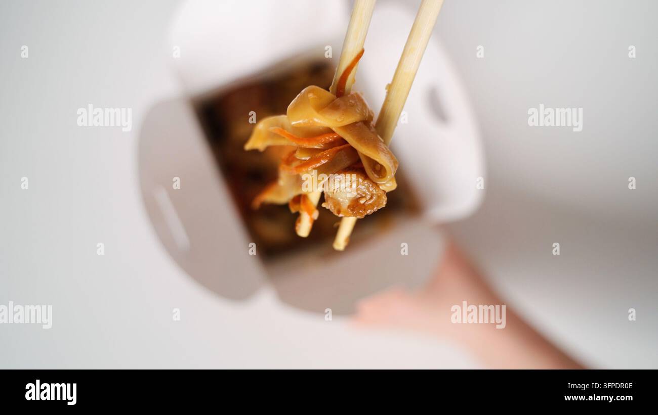 Persona che tiene le bacchette con noodle saltati e verdure, prende il cibo da una scatola da asporto bianca, pronta per mangiare Foto Stock