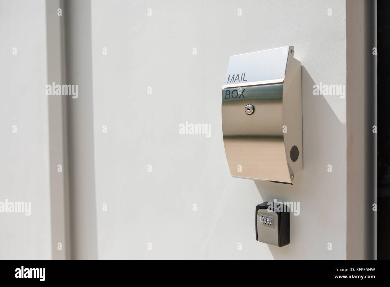 Cassetta delle poste moderna in acciaio inossidabile con scritta MAIL BOX montata su muro bianco, serratura a buca della chiave e cassaforte a combinazione sotto, luce del sole e ombra, grande c Foto Stock