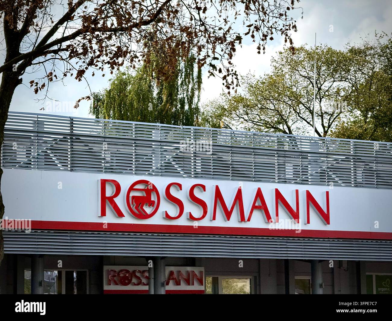 Una vista orizzontale della segnaletica della farmacia Rossmann con lettere rosse e logo a cavallo, montata su una facciata di edificio con persiane metalliche - Immagine stock catturata con smartphone