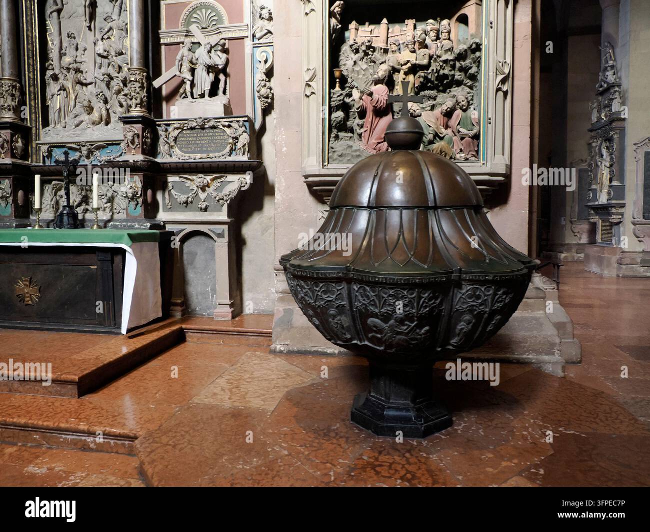 MAINZ, GERMANIA - 26 MAGGIO 2025: Un elegante fonte battesimale in bronzo con un coperchio a cupola e intagli a rilievo si trova all'interno della cattedrale di Magonza vicino a un elaborato Foto Stock
