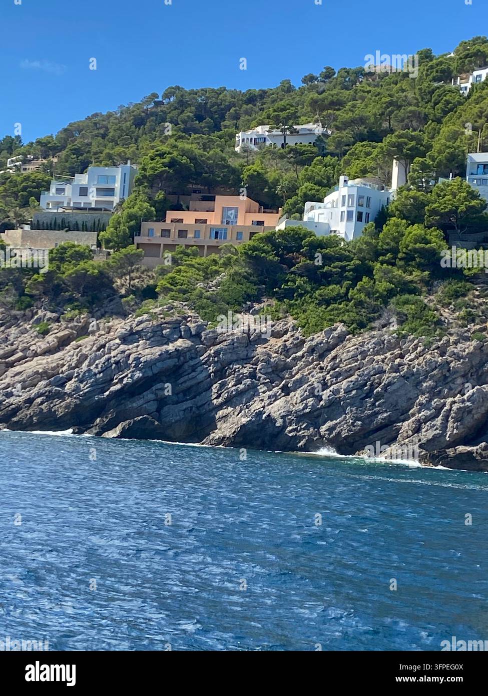 Ville di lusso moderne costruite su una ripida scogliera rocciosa circondata da una lussureggiante foresta di pini mediterranea, ripresa di tracciamento dall'acqua del mare. - Immagine stock catturata con smartphone