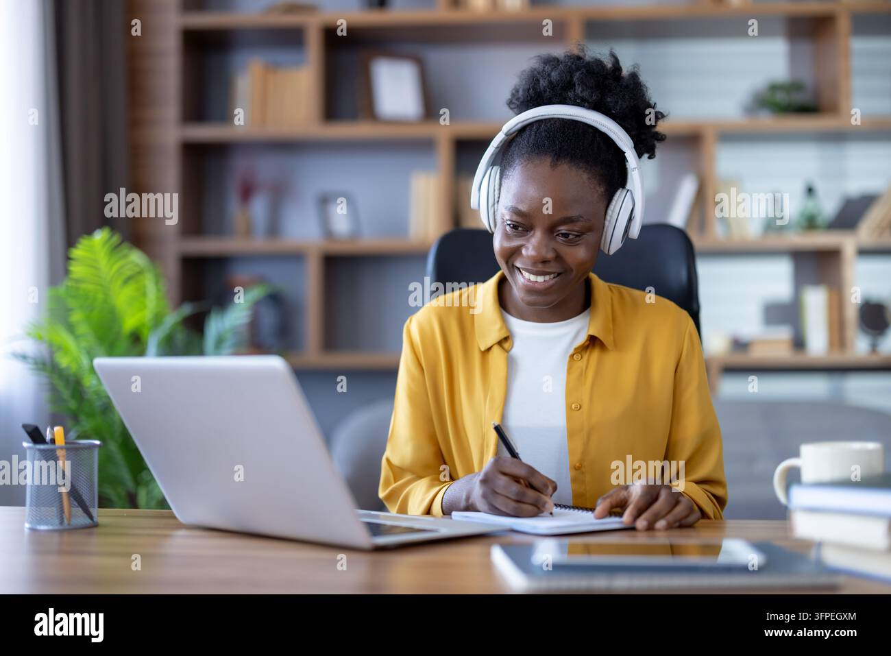 Donna sorridente con cuffie e che utilizza un laptop per l'apprendimento a distanza, il telelavoro o la partecipazione a un webinar, scrivendo in un quaderno alla scrivania dell'ufficio a casa Foto Stock