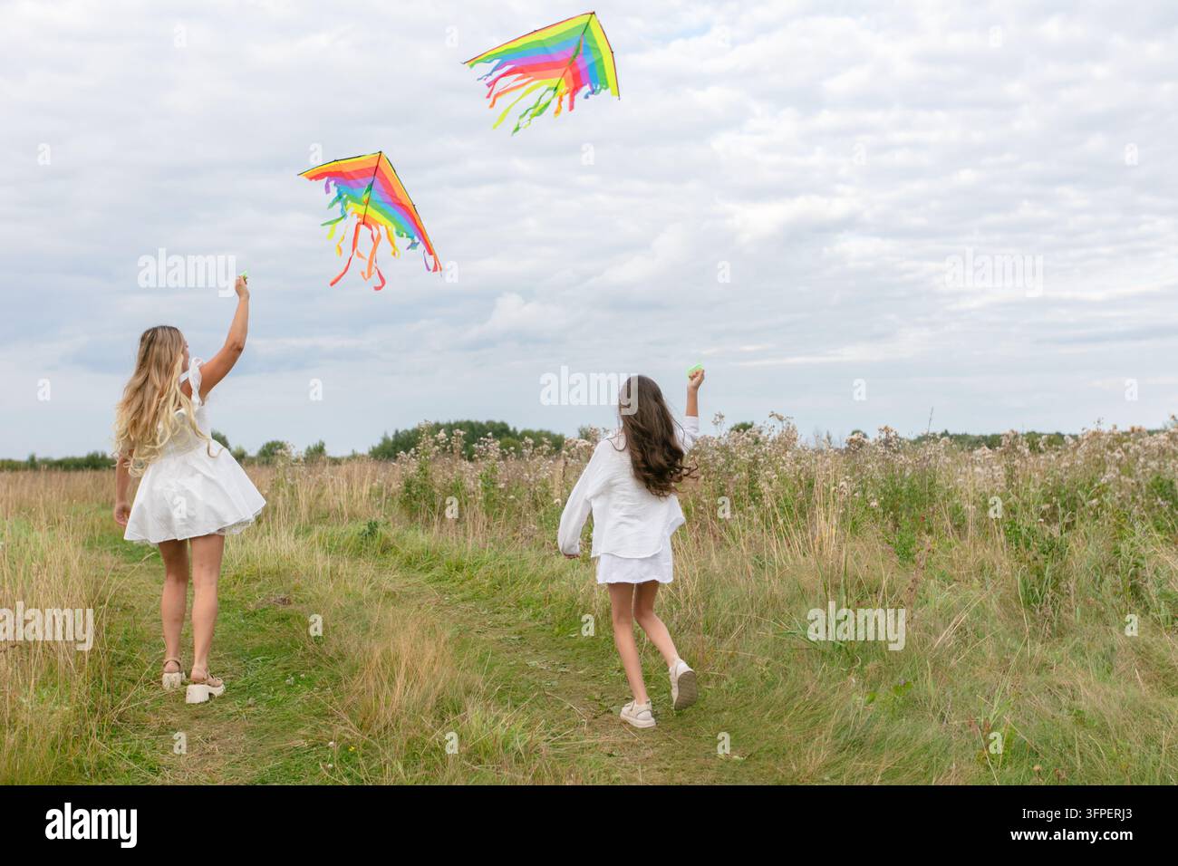 Mamma e figlia in abiti bianchi corrono attraverso un campo erboso, volano allegramente serpenti d'aria luminosi sullo sfondo di nuvole morbide. Celebrano Foto Stock