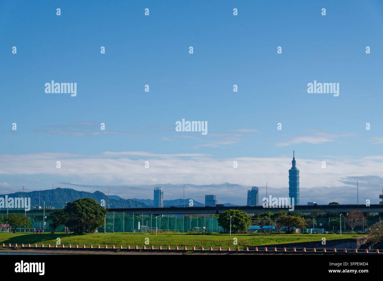 - 11 marzo 2026: Taipei 101 e altri grattacieli si ergono oltre un parco fluviale e una strada sopraelevata, con colline verdi e nuvole sottili sullo sfondo. Foto Stock