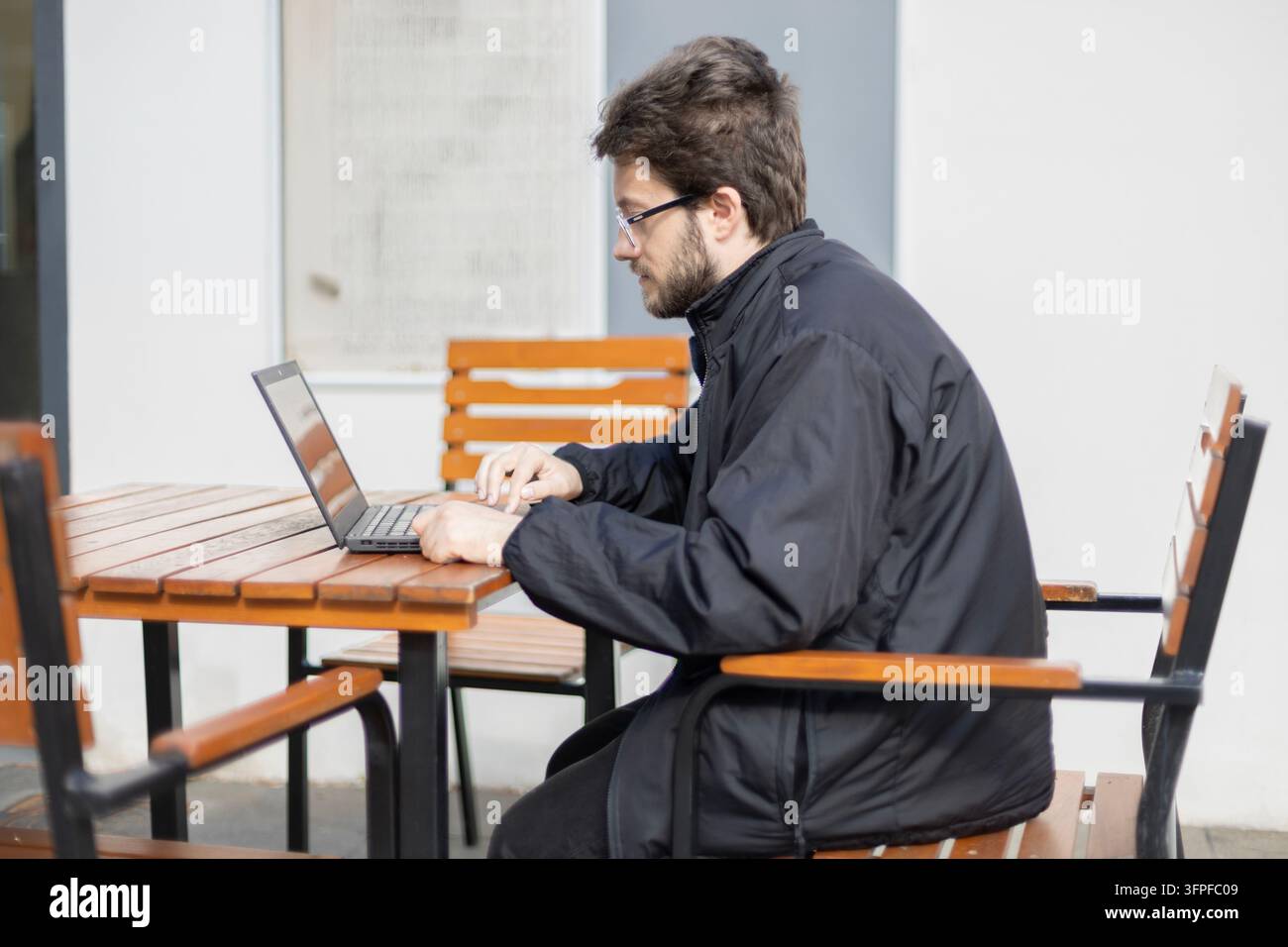 Un uomo con occhiali e giacca scura digita su un laptop mentre è seduto a un tavolo di legno in un'area esterna. La luce intensa del giorno illumina la s Foto Stock