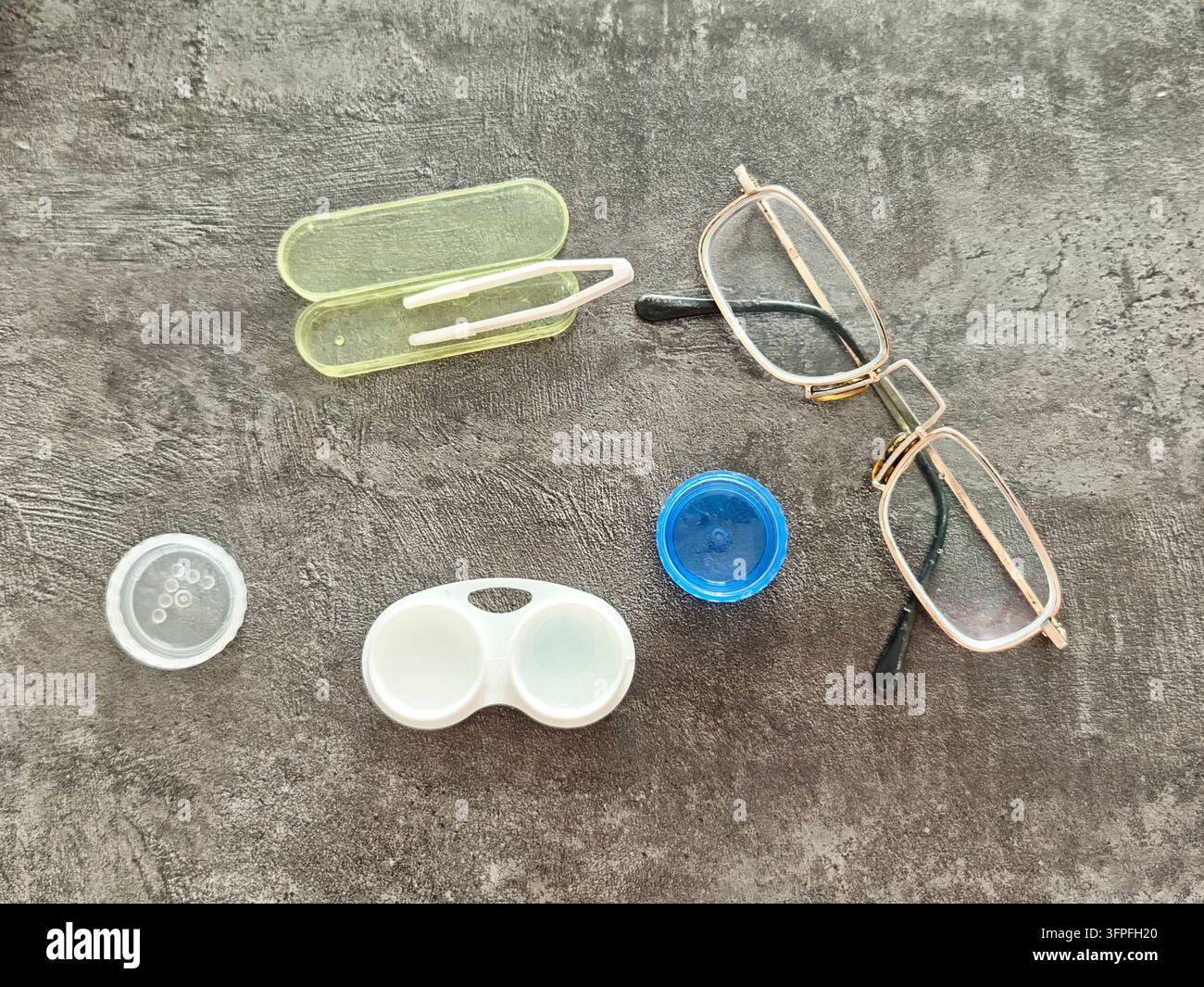 Articoli per la cura degli occhi sono disposti su una superficie grigia, comprese occhiali, un porta lenti a contatto e una pinzetta per maneggiare le lenti. Foto Stock