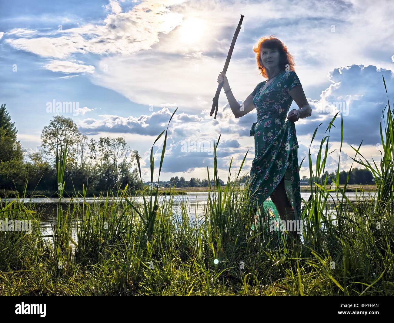 Una donna in abito a fiori si trova accanto a un corpo d'acqua calmo, tenendo un bastone mentre il sole tramonta dietro di lei, proiettando un bagliore caldo. Foto Stock