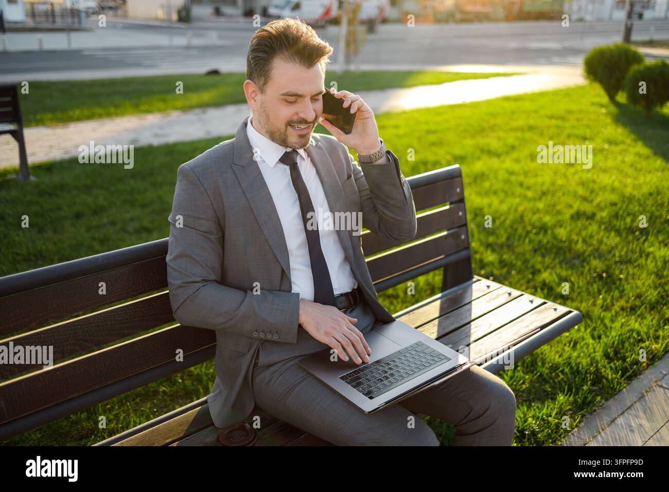 Un uomo in giacca e cravatta è seduto su una panchina con un laptop mentre parla al telefono. È giorno in un contesto di parco verdeggiante. Foto Stock