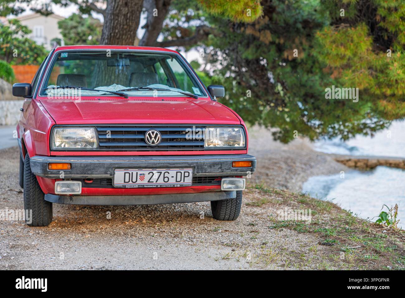 Orebic, Croazia – 19 maggio 2026: Volkswagen rosso parcheggiato su un sentiero costiero di ghiaia sotto i pini, vista frontale con paraurti e area della targa visibili Foto Stock